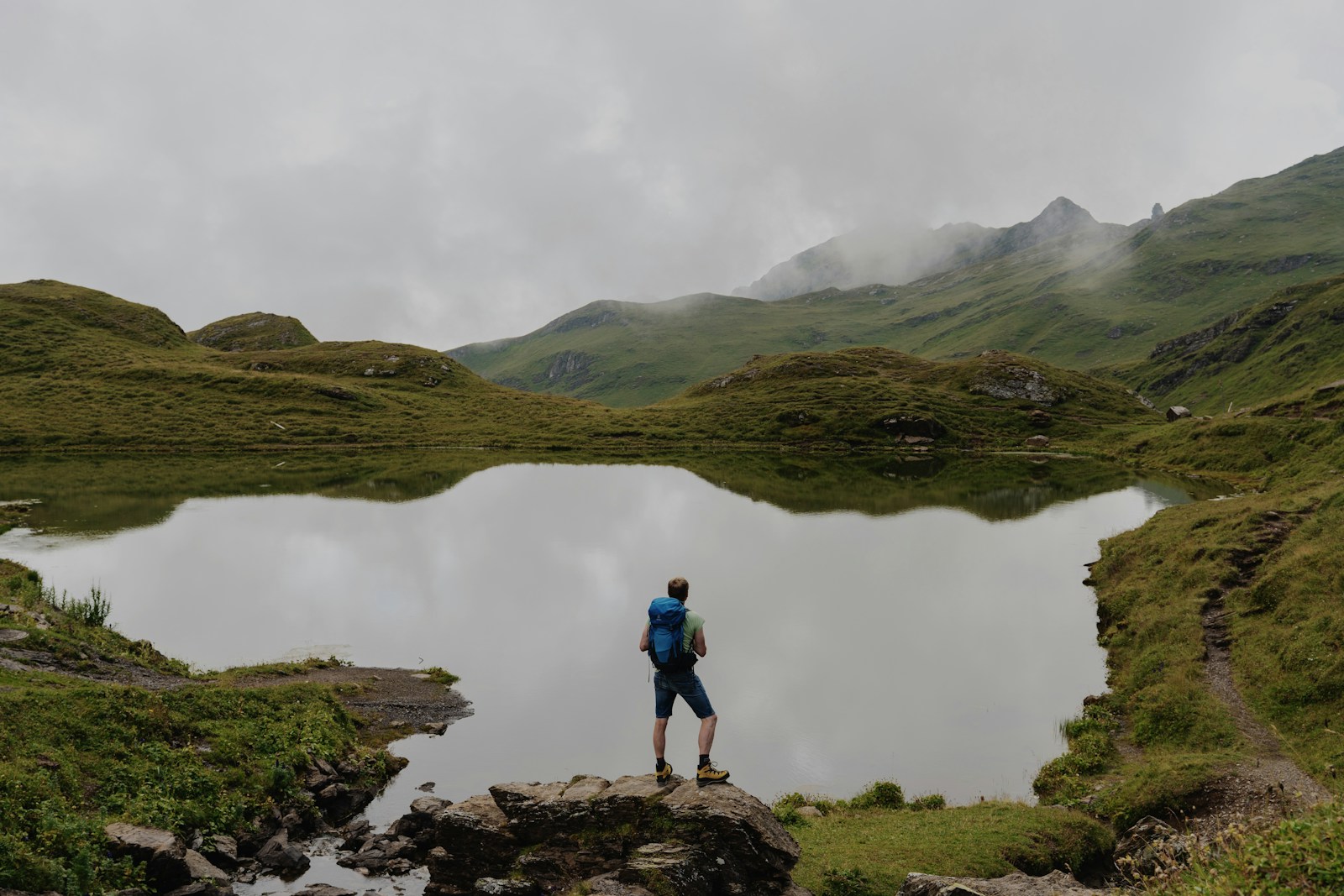 Hiker stands by a calm mountain lake on a cloudy day.