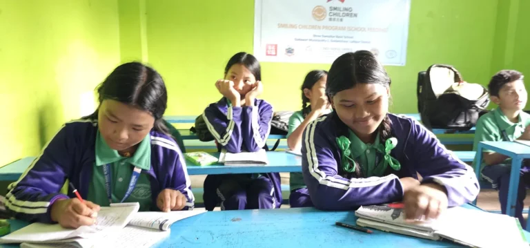 बैजनी ज्याकेट लगाएका विद्यार्थीहरू निलो डेस्कमा कापीमा लेख्दै, भित्तामा स्माइलिङ चिल्ड्रेन कार्यक्रमको ब्यानर