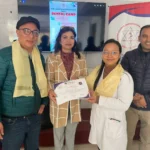 दन्त शिविरको पोस्टर देखिएको स्क्रिन र AAAK ब्यानरअगाडि प्रमाणपत्र समातेका चार जना