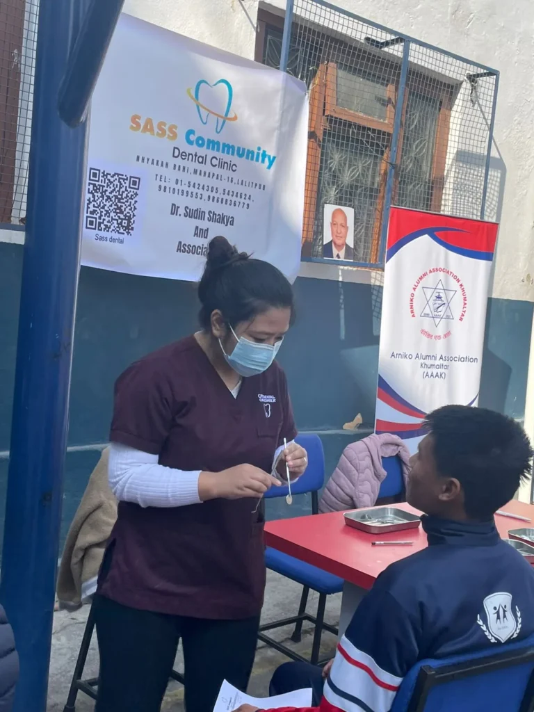 बसेका विद्यार्थीछेउ उपकरण तयार गर्दै दन्त स्वास्थ्यकर्मी, पछाडि क्लिनिक र AAAK का ब्यानर