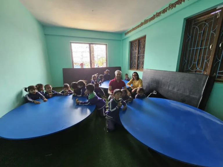 नयाँ रंग लगाइएको कक्षामा गोलो निलो टेबल वरिपरि बसेका साना बालबालिका