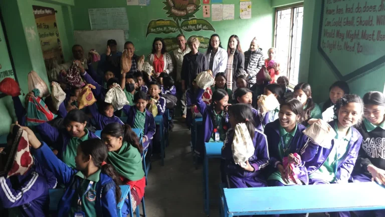 Projects रंगीन कपडाका झोला उचालेर मुस्कुराइरहेका विद्यार्थीले भरिएको कक्षा, पछाडि उभिएका अतिथिहरू