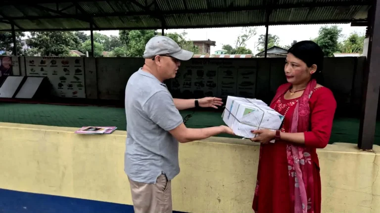छाना भएको विद्यालयको खेल्ने ठाउँमा रातो कुर्ता लगाएकी शिक्षिकालाई कापीको बन्डल हस्तान्तरण गर्दै टोपी लगाएका व्यक्ति