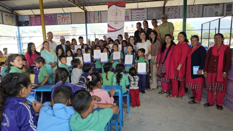 छाना भएको विद्यालय आँगनमा AAAK ब्यानरछेउ प्रमाणपत्र लिएका हरियो र बैजनी पोसाकका विद्यार्थी, अभिभावक, शिक्षक र AAAK सदस्य