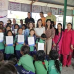 AAAK ब्यानरछेउ कदर पत्र लिएका हरियो पोसाकका विद्यार्थी र पछाडि उभिएका अतिथि