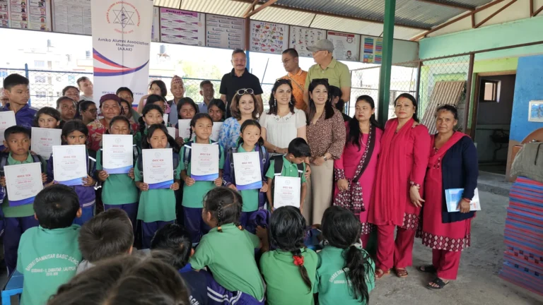 AAAK ब्यानरछेउ कदर पत्र लिएका हरियो पोसाकका विद्यार्थी र पछाडि उभिएका अतिथि