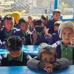 जाडोको पोसाकमा निलो डेस्कमा बसेका साना विद्यार्थीहरू, केही क्यामेरातिर मुस्कुराउँदै