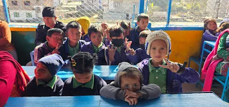 जाडोको पोसाकमा निलो डेस्कमा बसेका साना विद्यार्थीहरू, केही क्यामेरातिर मुस्कुराउँदै